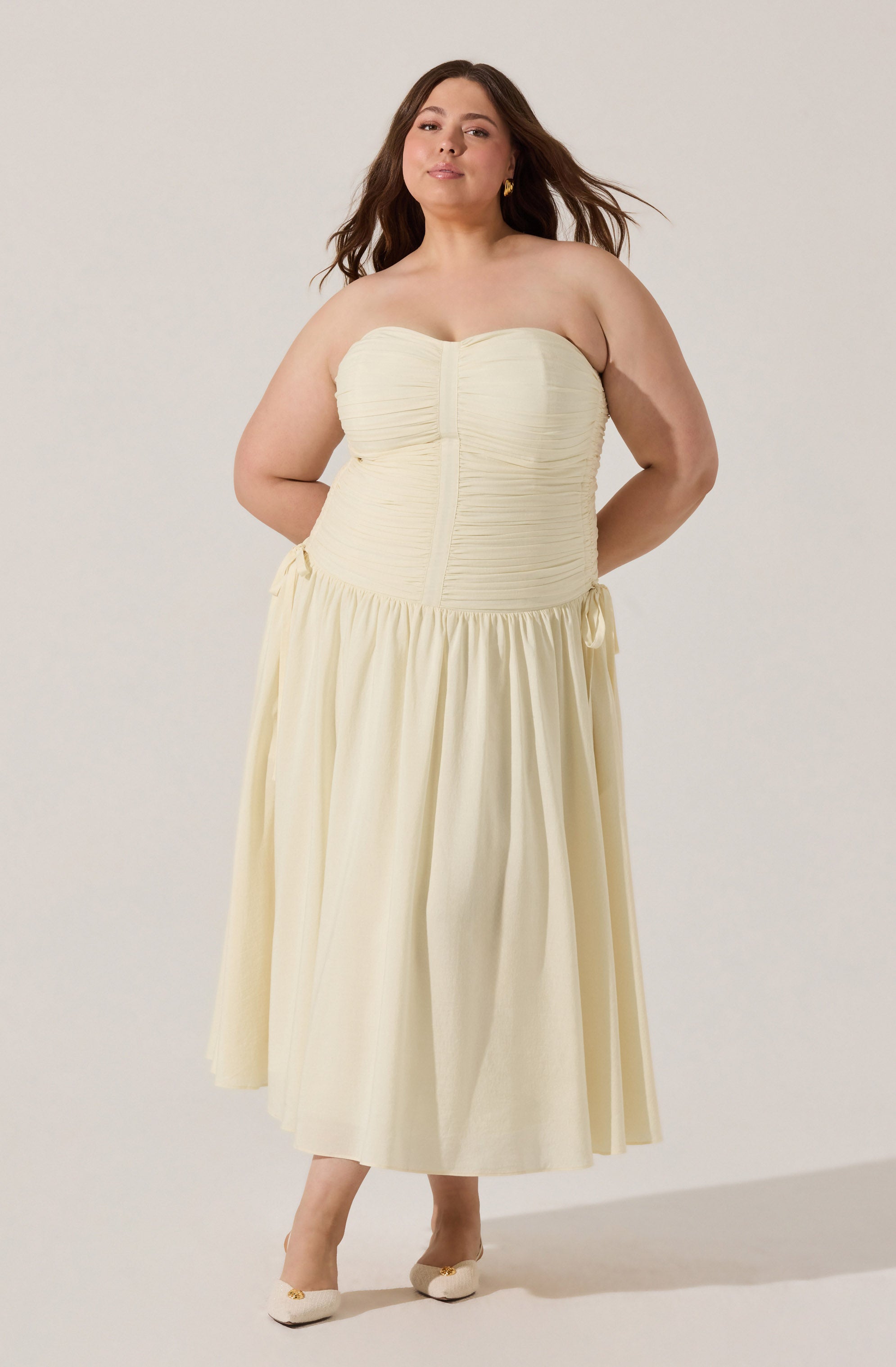 A woman wearing a strapless, cream-colored dress with ruched detailing along the bodice and a gathered, flowing skirt that falls to her ankles. She has on matching cream shoes with a decorative element on the front.