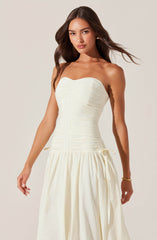 A woman is wearing a strapless, fitted, cream-colored dress with horizontal ruching on the bodice. The dress features a seam down the center and a gathered, flowing skirt with a tie at the waist.