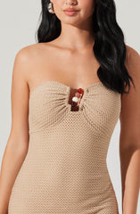 A woman is wearing a strapless, beige textured dress that features a gathered bust with a small opening in the center, decorated with large pearl and marble beads. She has long, dark, wavy hair and is accessorized with large, shiny hoop earrings.