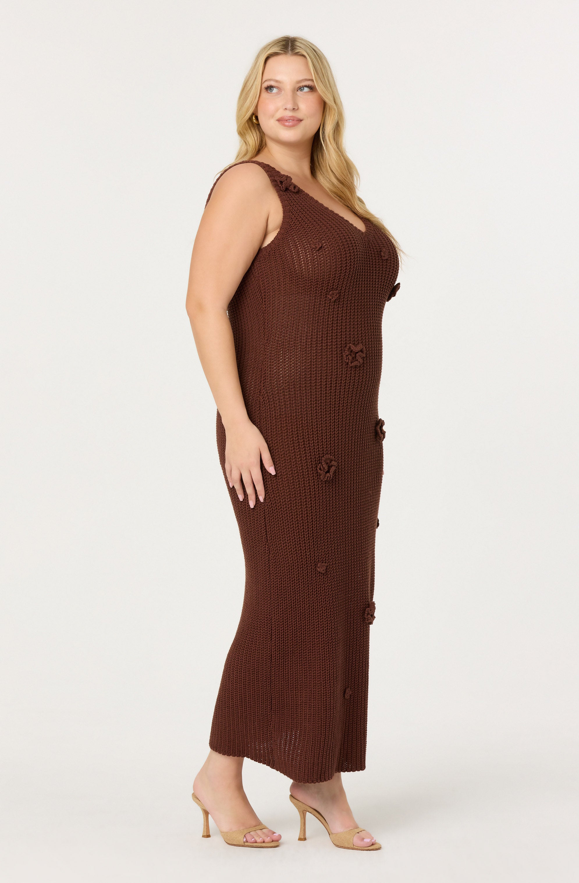 A woman is wearing a sleeveless, ankle-length dress made of a chunky, dark brown crochet material. The dress features decorative crochet flowers along the front and also has ruffle details on the shoulder straps. She is wearing nude, open-toe high heels.