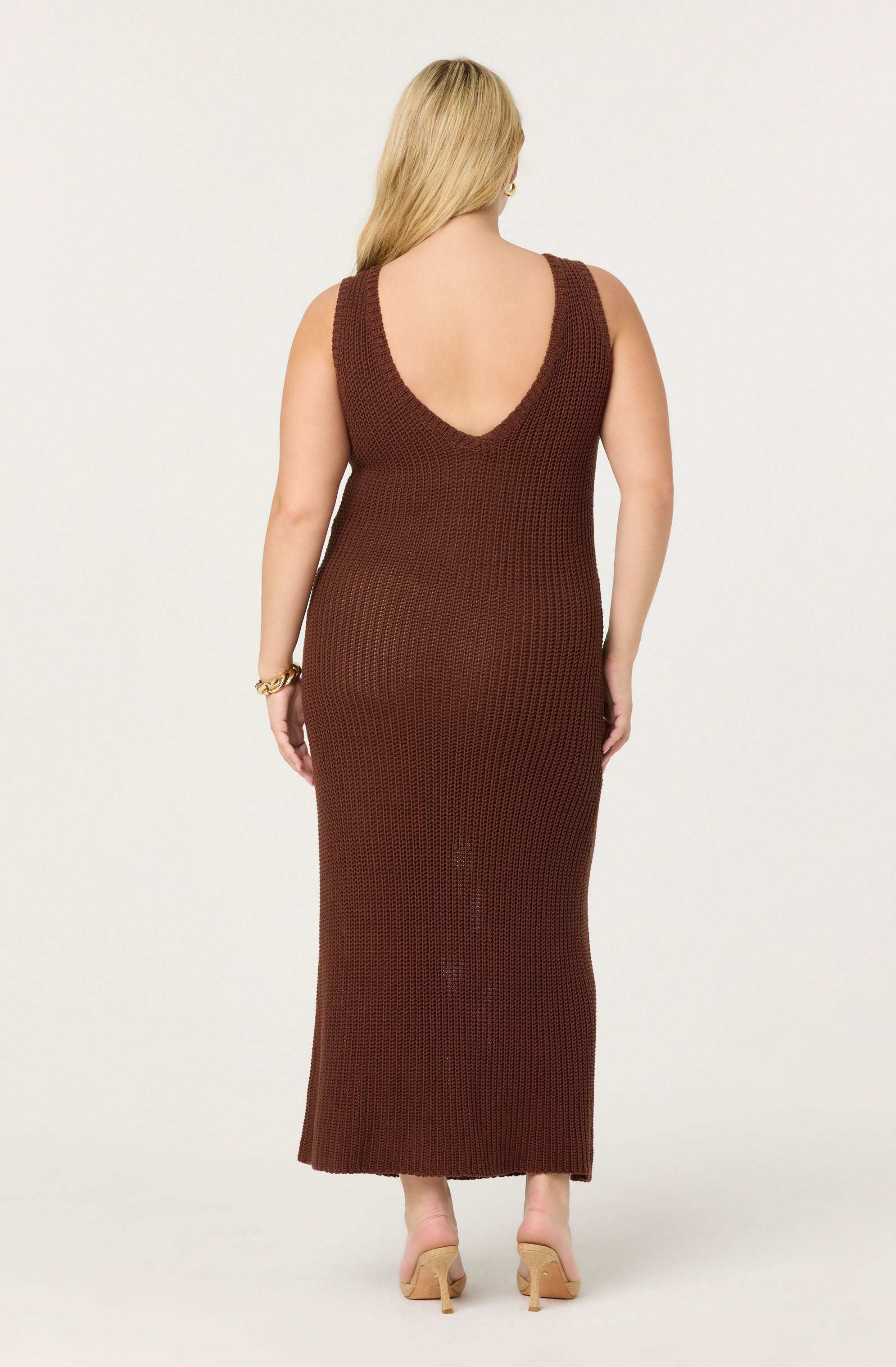 A woman is wearing a sleeveless, ankle-length, knitted dress in a dark brown color. The dress has a deep U-shaped neckline at the back. She is also wearing nude-colored high-heeled shoes and a gold bracelet on her left wrist.