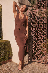 A woman is wearing a sleeveless, ankle-length, brown knit dress with small distressed holes. She accessorizes with a large circular pendant necklace and strappy heels.