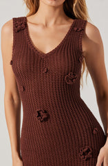A woman is wearing a sleeveless, V-neck, knitted dress in a rich brown color. The dress features textured stitch patterns and decorative, three-dimensional floral appliqués scattered across the fabric. The shoulders have small knitted bows as embellishments.