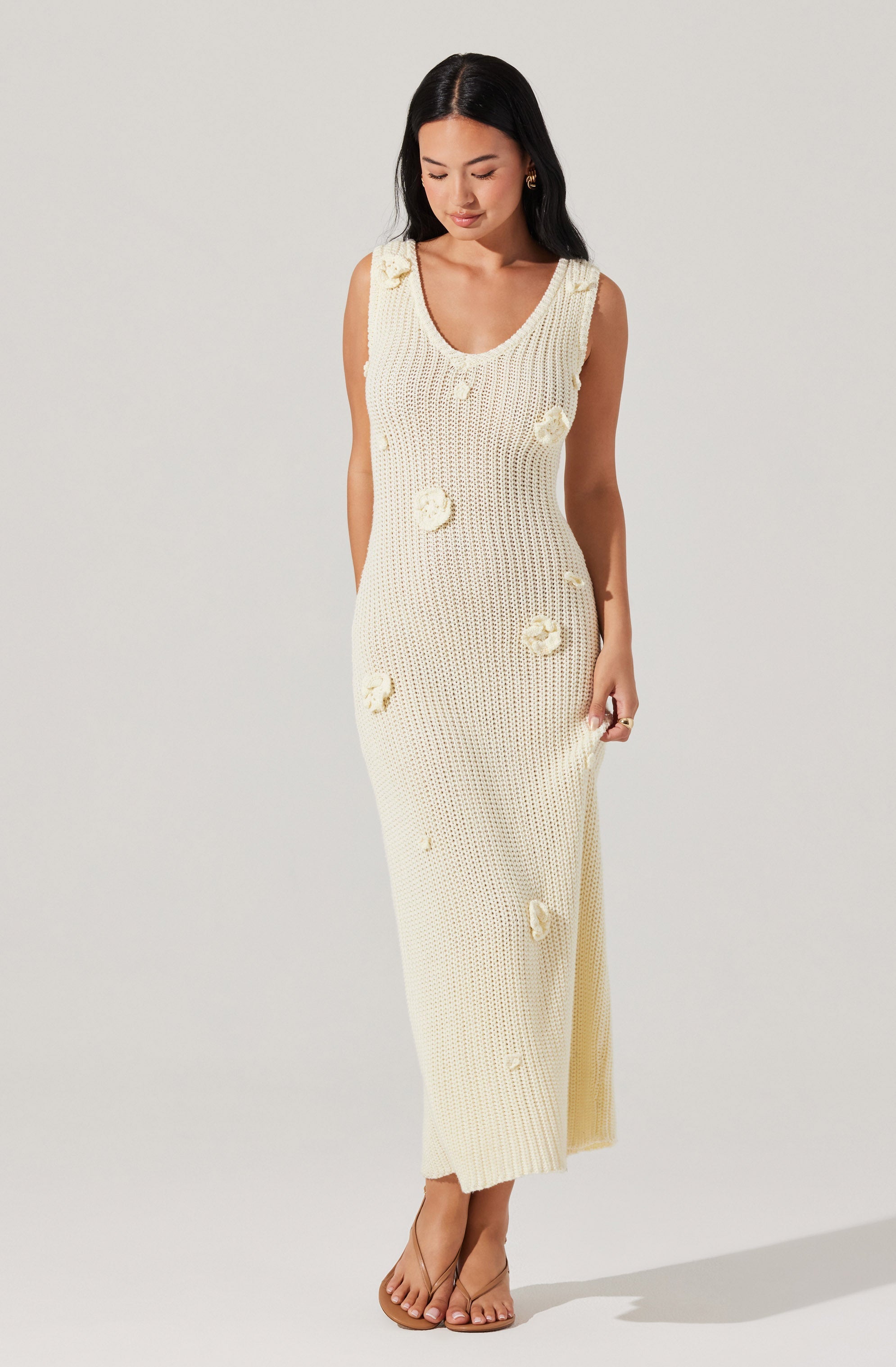 A woman is wearing a sleeveless, cream-colored, knit maxi dress with a scoop neckline. The dress features decorative flower appliqués scattered across the fabric. She is also wearing clear, open-toe high heels.
