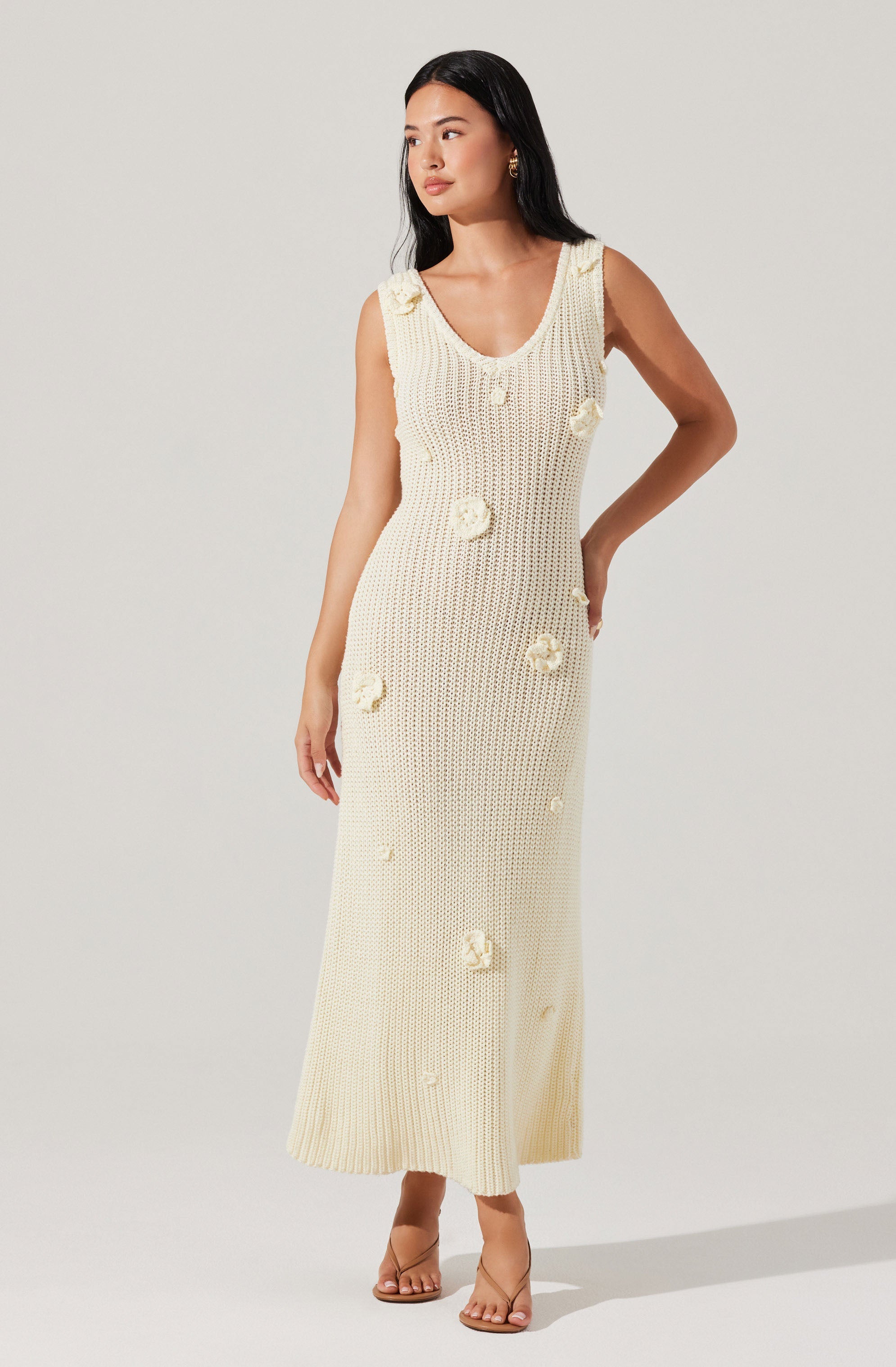 A woman is wearing a sleeveless, cream-colored, knit dress that extends to her ankles. The dress features a scooped neckline and is decorated with textured floral appliqués scattered throughout. She is also wearing transparent-strap nude heels.