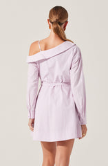 A woman is wearing a light pink shirt dress with long sleeves and buttoned cuffs. The dress has an off-the-shoulder design, exposing one shoulder with a strap, and features a collar that falls off one side. The dress is belted at the waist, creating a cinched look, and has a short hemline above the knees.