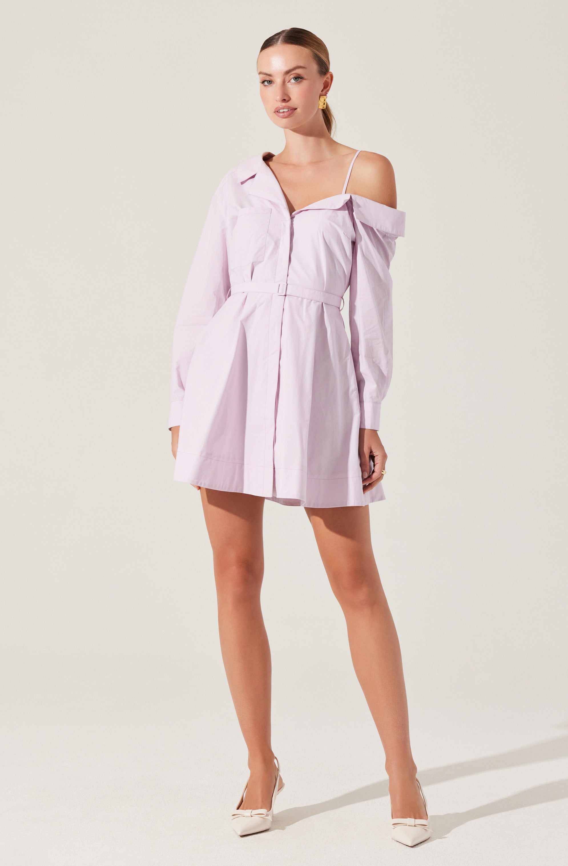 A woman is wearing a light pink, off-the-shoulder shirt dress with a collar and long sleeves. The dress has a gathered waist, button details, and a slightly flared skirt. She is also wearing a white spaghetti strap top underneath, with pointed white heeled shoes.