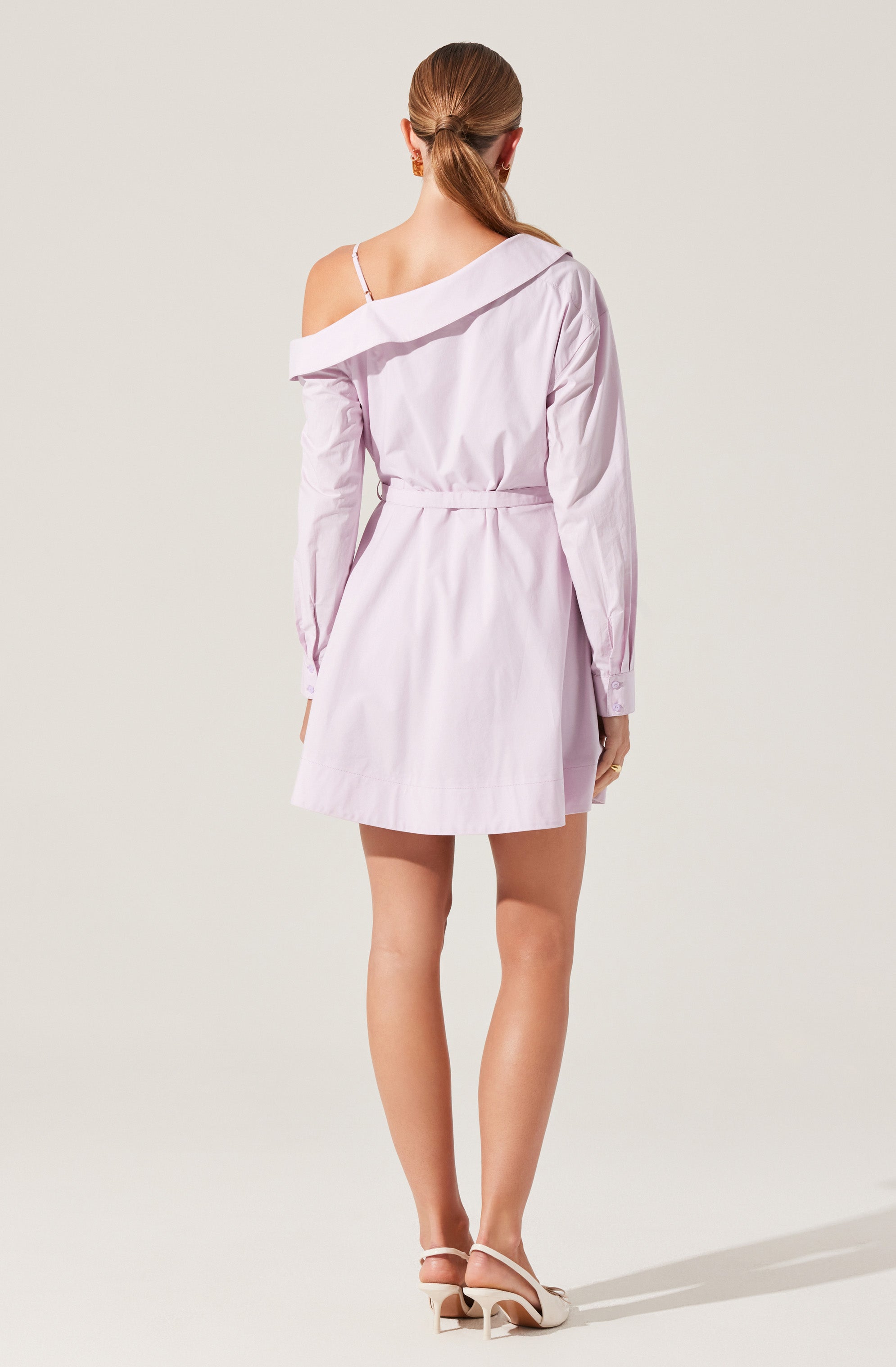 A woman is wearing a light purple, off-the-shoulder shirt dress with long sleeves. The dress has a loose fit with a waist tie and features a single strap detail on her left shoulder. She is also wearing light-colored, open-toe high heels with ankle straps.