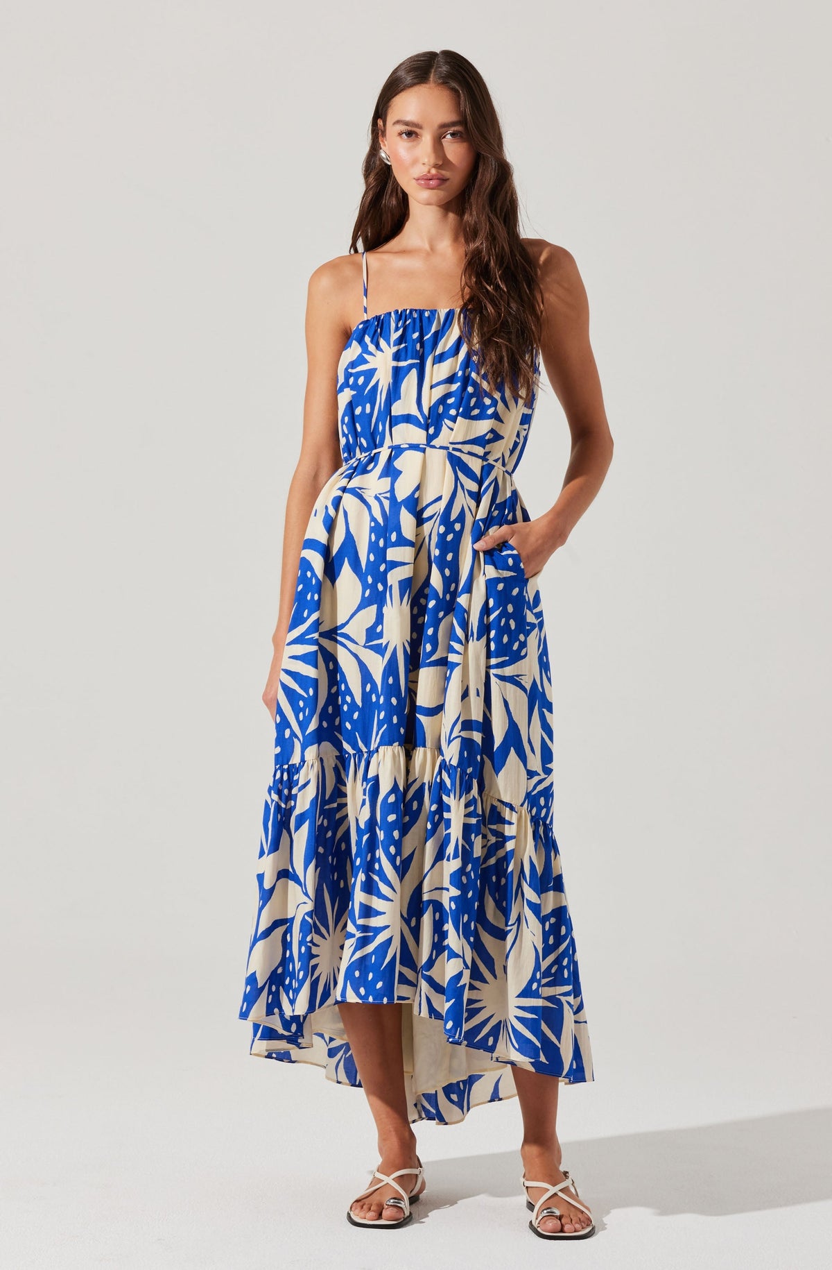 A woman is wearing a sleeveless, ankle-length dress with thin spaghetti straps. The dress features a bold, abstract tropical print in shades of blue and cream. It has a loose, flowing fit with a gathered bodice and a waistband that ties at the side. She is also wearing open-toe, strappy sandals in a light color.