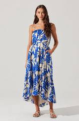 A woman is wearing a sleeveless, ankle-length dress with thin spaghetti straps. The dress features a bold, abstract tropical print in shades of blue and cream. It has a loose, flowing fit with a gathered bodice and a waistband that ties at the side. She is also wearing open-toe, strappy sandals in a light color.
