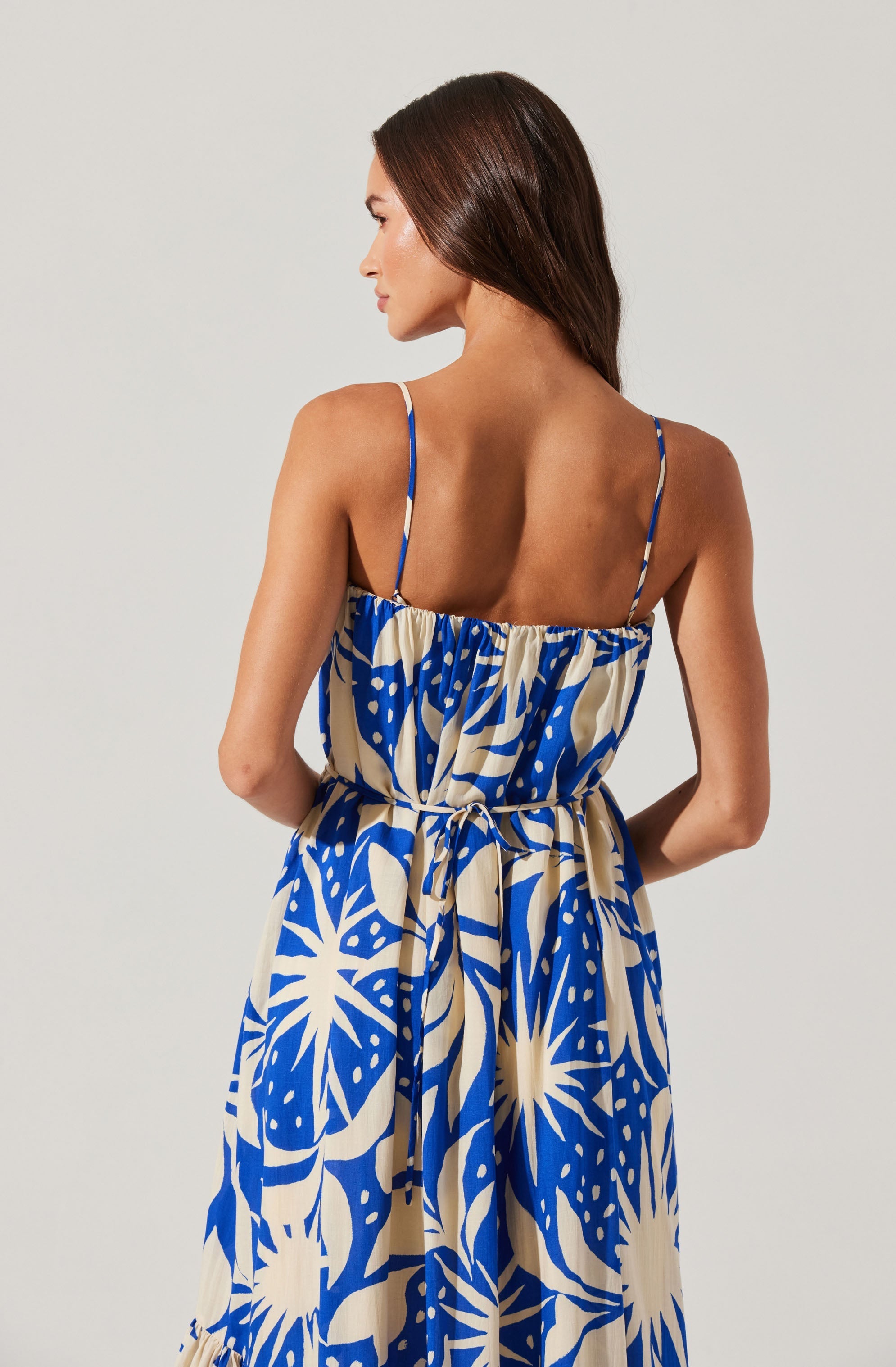 A woman is wearing a sleeveless dress with thin spaghetti straps. The dress features a bold, abstract pattern in blue and beige, with large leaf and sunburst shapes. The dress has a gathered waistline with a tie, creating a casual and flowing silhouette. The woman has long, brown hair and is posed with her back to the camera, standing against a plain, light-colored background.