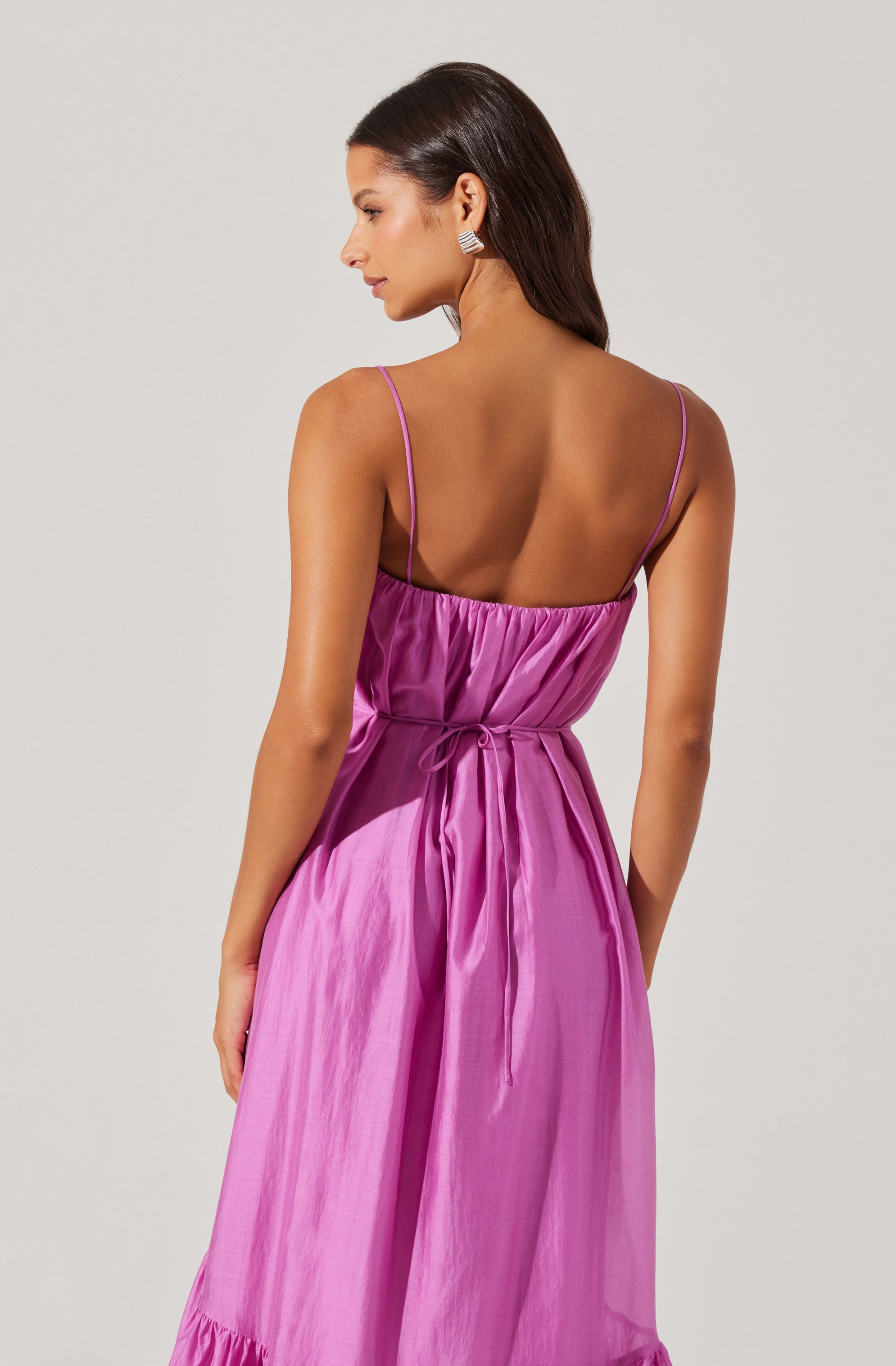 A woman is wearing a sleeveless, pink satin dress with thin spaghetti straps. The dress has a gathered bodice with a drawstring tie at the waist and a ruffled hem at the bottom. She is also wearing silver statement earrings.