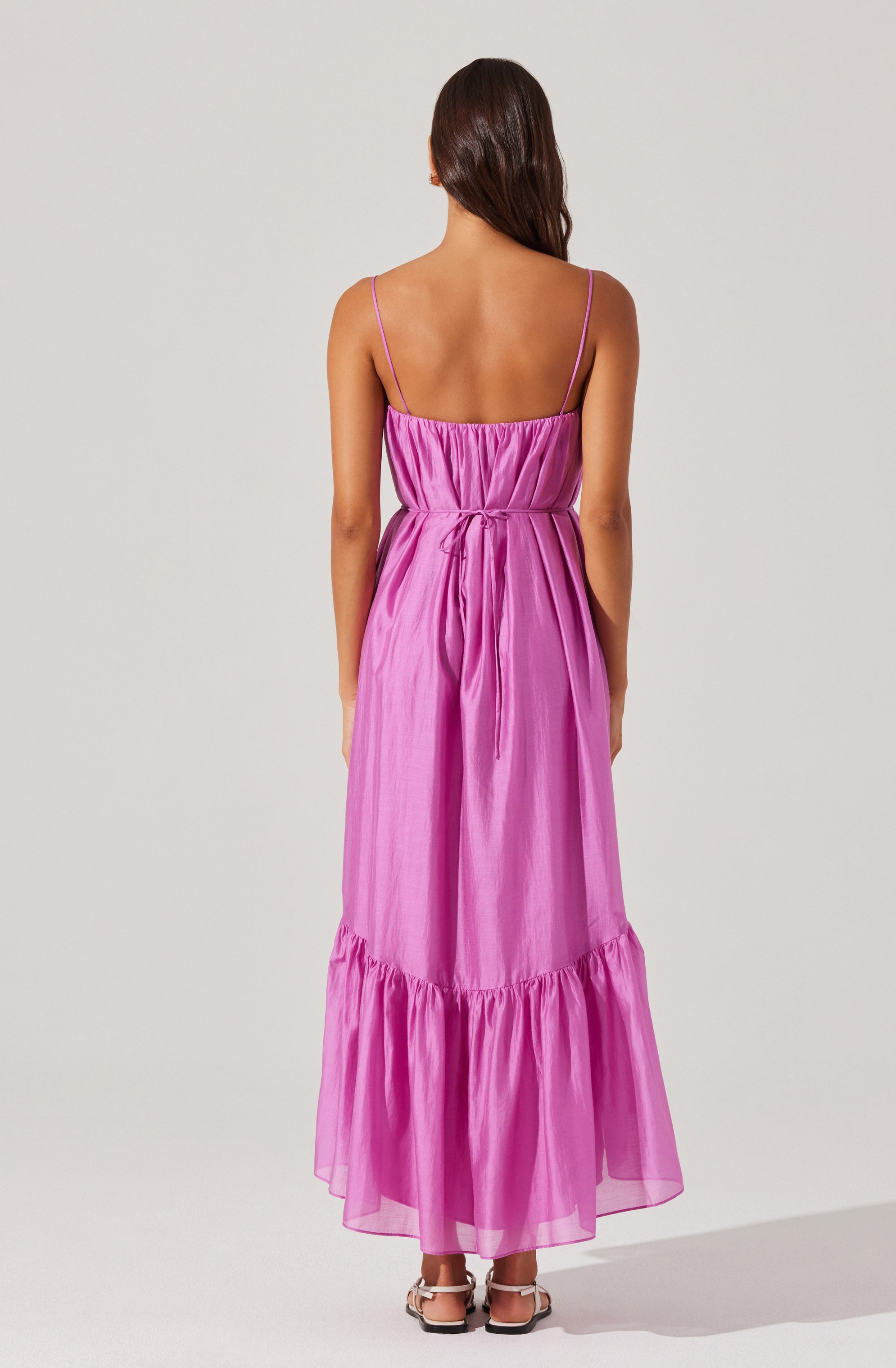 A woman is wearing a long, pink, sleeveless dress with thin shoulder straps. The dress has a gathered bodice and a slight cinch at the waist, with a fabric tie. The skirt is flowy and features a ruffled hem at the bottom.