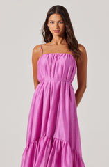 A woman is wearing a bright pink, sleeveless, ankle-length dress with thin spaghetti straps. The dress has a gathered, elasticized bodice and a tiered skirt with ruffle details at the hem. The fabric appears shiny and lightweight.