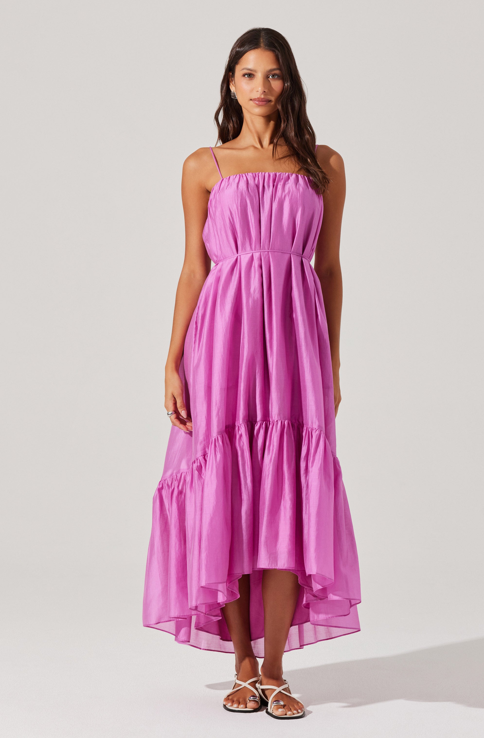 A woman is wearing a sleeveless, floor-length dress with thin spaghetti straps. The dress features a gathered, elasticized bodice and a flowing, A-line skirt with a ruffled hem that is slightly shorter in the front and longer in the back. The fabric of the dress appears to be lightweight and shiny, with a vibrant pink color. She completes the look with white, strappy sandals.