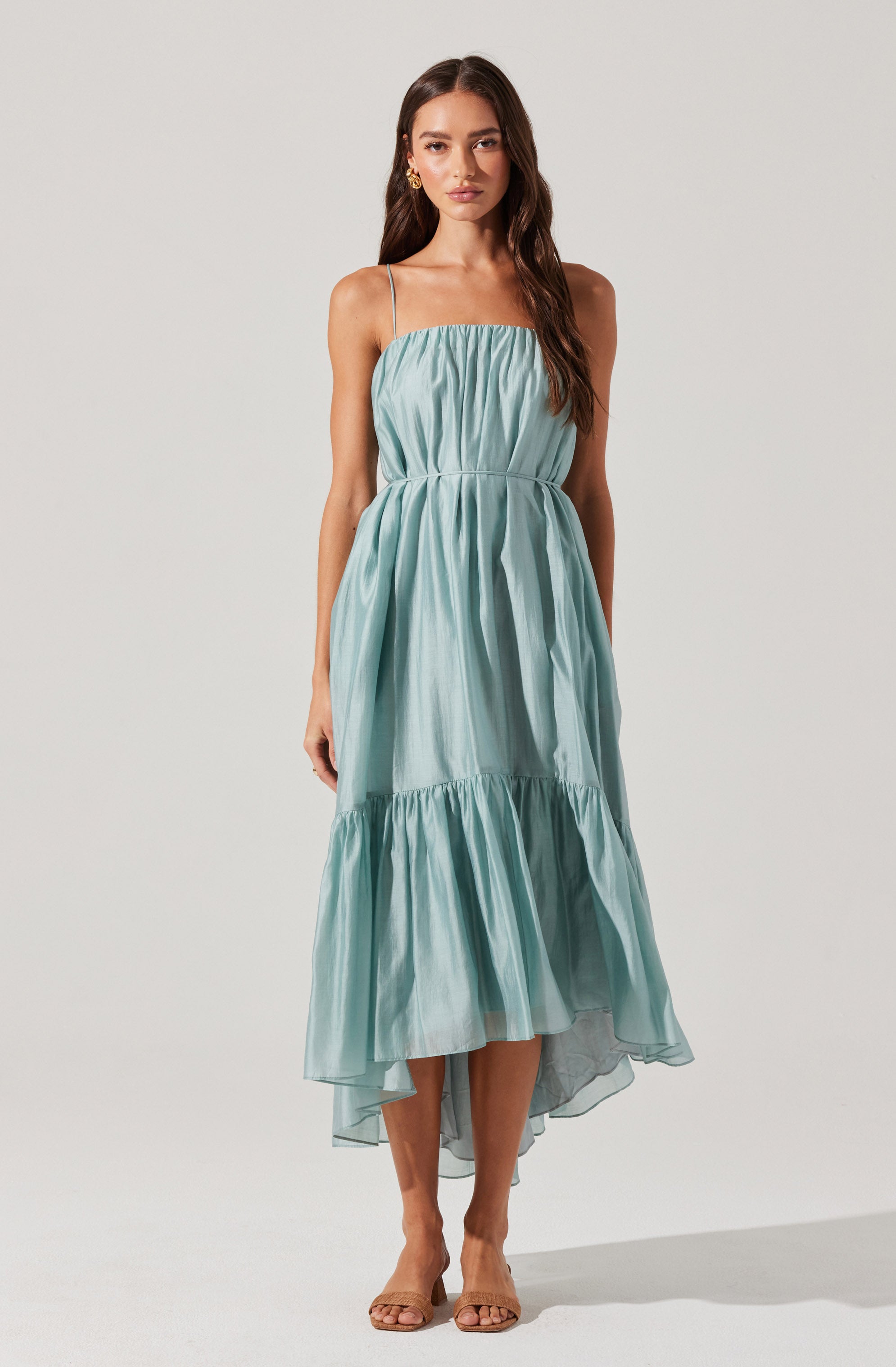A woman is wearing a sleeveless, light blue maxi dress with thin spaghetti straps. The dress has a gathered, ruched bodice and a loose-fitting skirt with a ruffled hem that is slightly shorter in the front and longer in the back. She is also wearing open-toed, nude-colored high heels and gold hoop earrings.