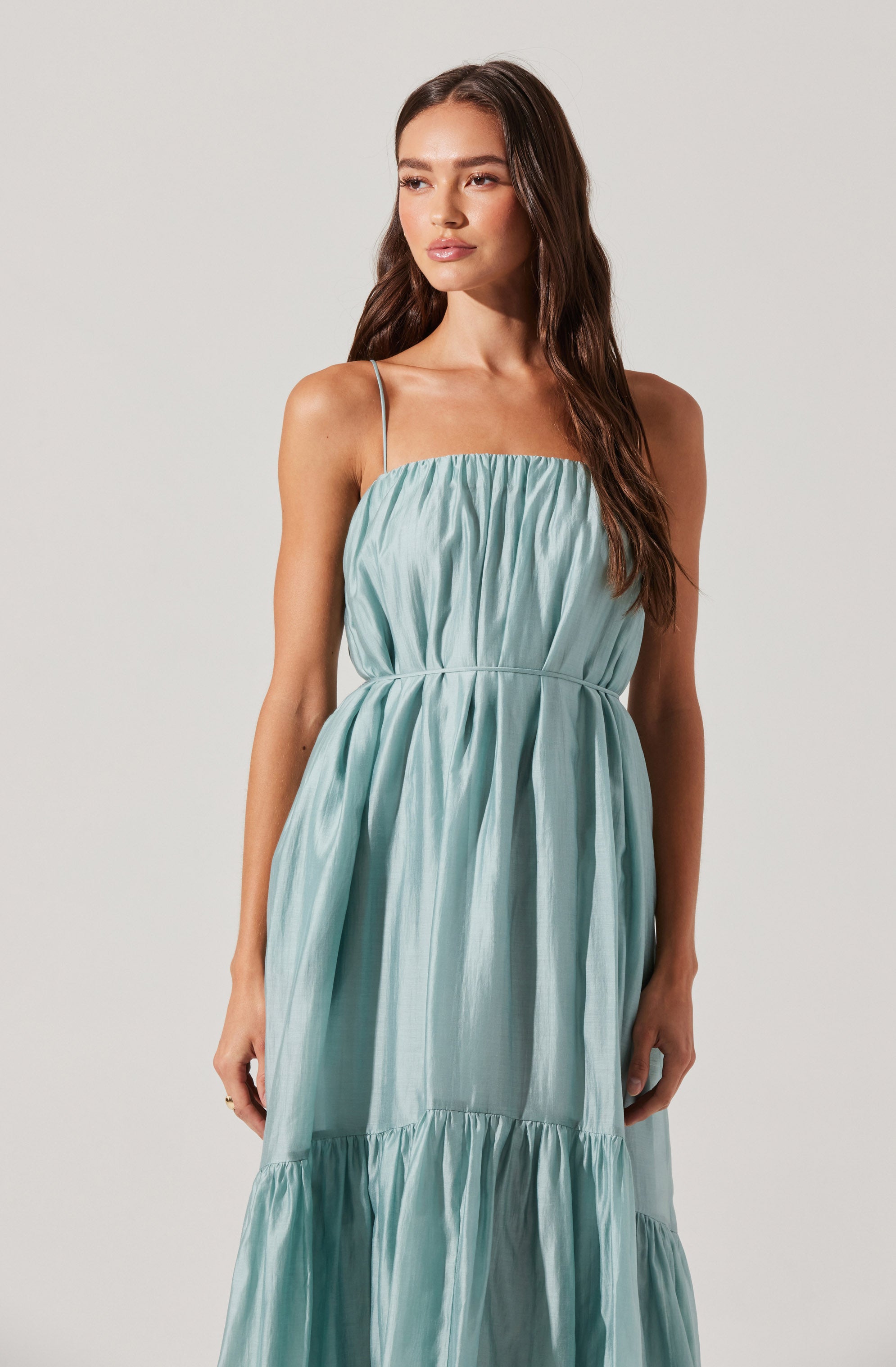 A woman is wearing a sleeveless, light blue dress with spaghetti straps. The dress features a gathered, ruched bodice and a waistline seam, with a flowing skirt that has a tiered design with gathered fabric.