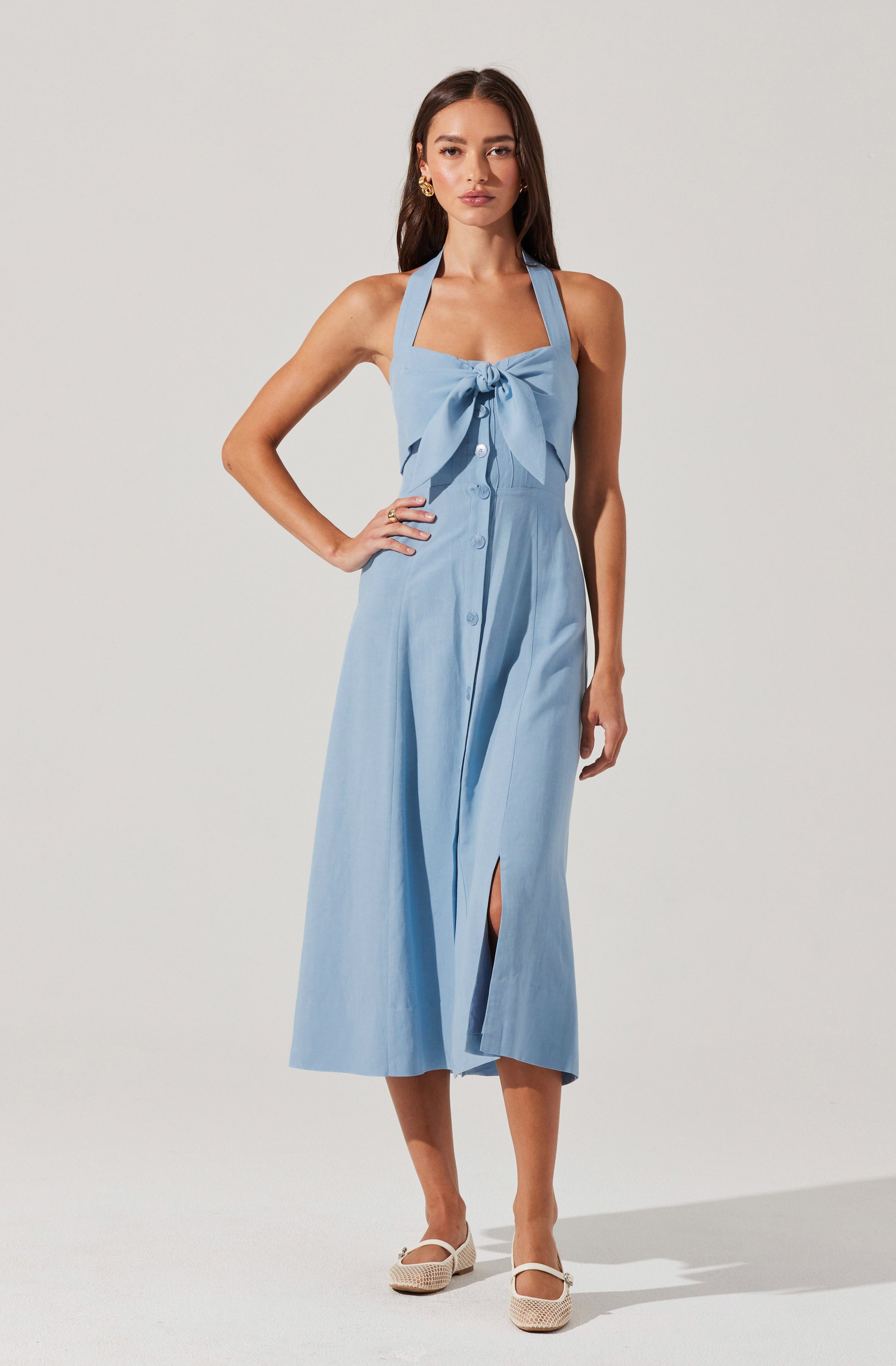 A woman is wearing a light blue sleeveless dress with wide straps that form a halter neckline. The dress features a large bow detail on the chest, a button-down front, and a slit in the skirt. She is also wearing white, woven, closed-toe shoes.