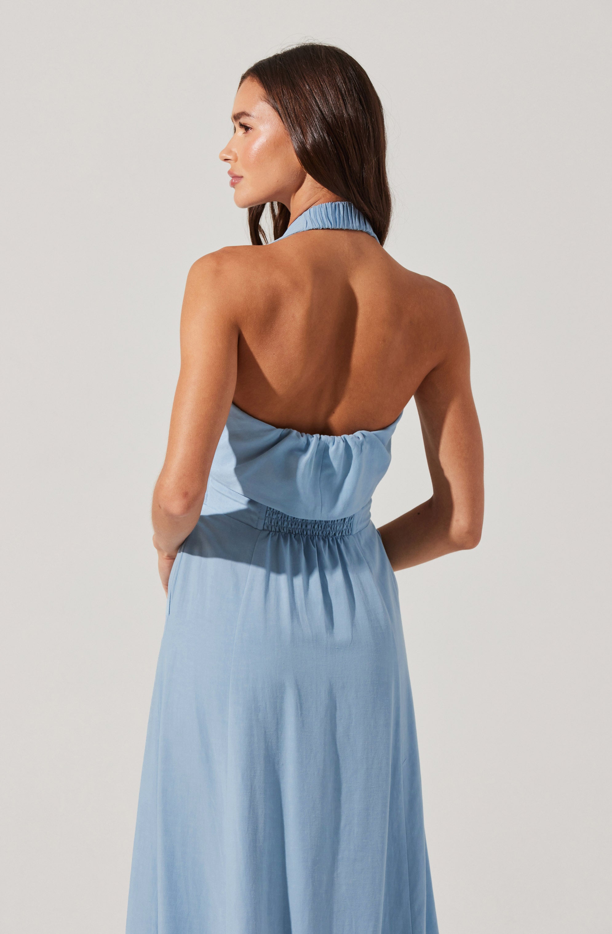 A woman is wearing a light blue, sleeveless dress. The dress has a high, ruffled collar around the neck and an open back with gathered fabric below the neckline. The waist is cinched with elastic, and the skirt is long and flowing, made of a lightweight fabric.