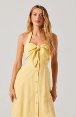 A woman is wearing a sleeveless, light yellow dress with a large bow on the front. The dress features a row of buttons running down the center, and the top part has a halter neckline with a tied bow. The fabric appears lightweight and summery.