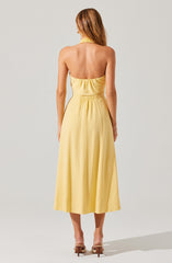 A woman is wearing a sleeveless, yellow halter-neck dress with an elastic waistband. The dress has a gathered bodice and a long, flowing skirt that reaches mid-calf. She is standing with her back to the camera, and her hair is styled in loose waves.