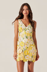 A woman is wearing a sleeveless dress with a V-neckline. The dress features a floral pattern with large yellow and white flowers and green leaves, and the fabric has a textured, embroidered appearance. The hem of the dress is above the knee.