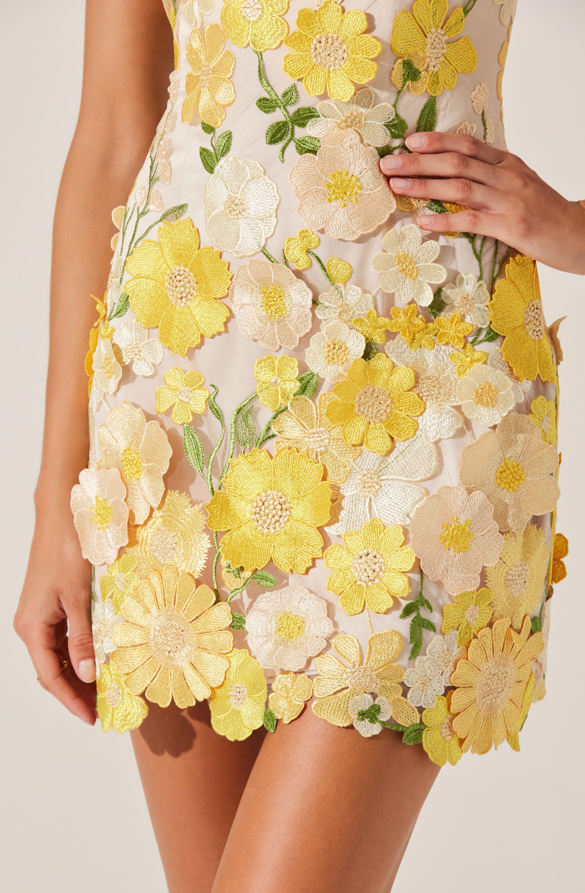 A close-up of a woman's dress featuring an intricate floral embroidery pattern with yellow, cream, and pastel pink flowers, along with green leaves. The dress is made of a sheer, light fabric with three-dimensional embroidered flowers covering the entire front. The woman's right hand is resting on her waist, while her left hand hangs down by her side.