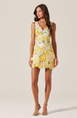 The dress is sleeveless with a V-neckline and features a floral pattern with yellow and white flowers with green leaves. It has a fitted waist and a short hemline, with the floral embroidery extending throughout the entire dress, including the hem. The overall look is feminine and summery.