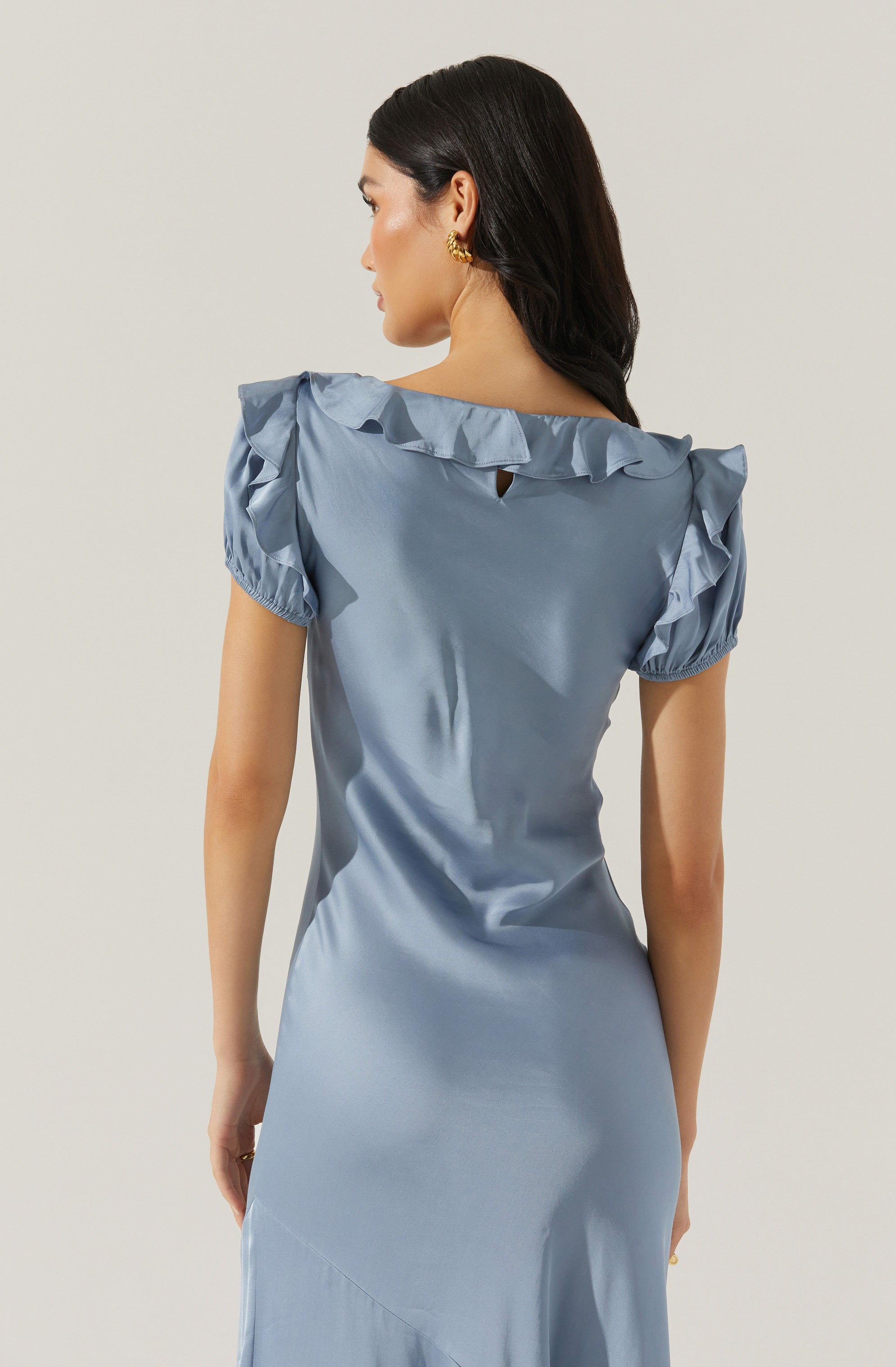 A woman is wearing a light blue satin dress with ruffled cap sleeves. The dress has a slight sheen and features a small keyhole opening at the back neckline. She has dark, wavy hair and is accessorized with gold earrings.