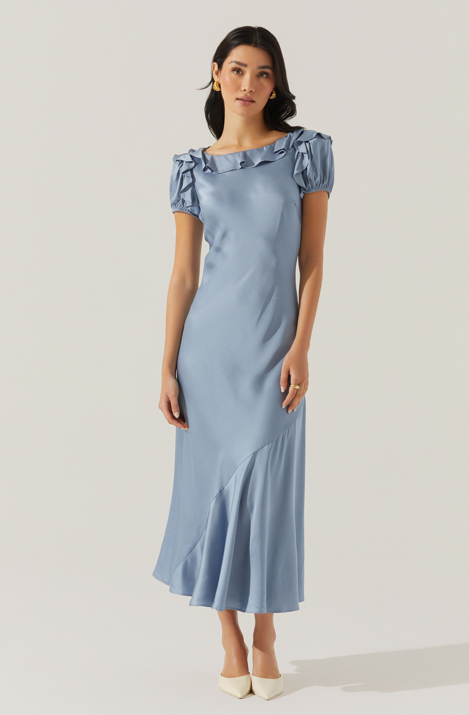 A woman is wearing a light blue, satin-like dress with short puffed sleeves that have ruffled detailing along the neckline and shoulders. The dress is fitted at the waist and flows down to a midi length with a subtle draped ruffle detail at the hem. She is also wearing white high heels and gold earrings.