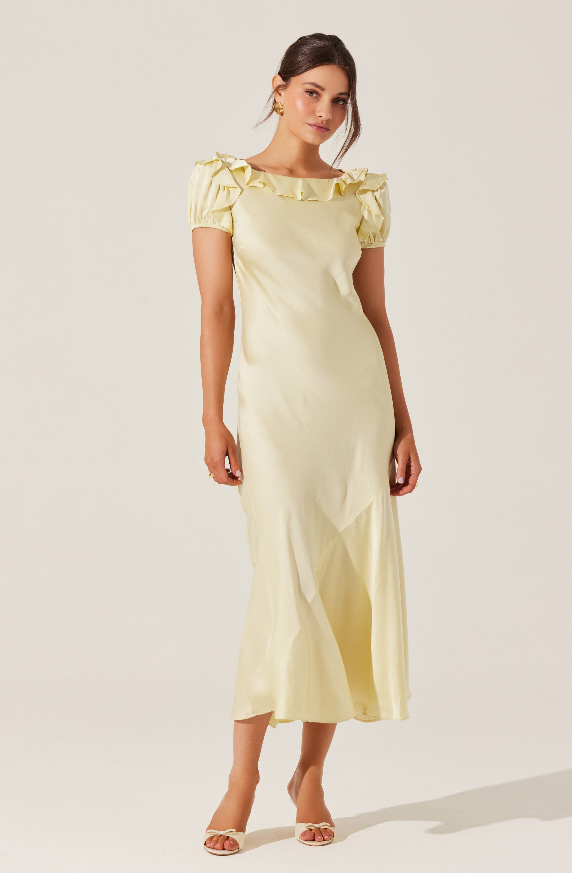 A woman is wearing a light yellow, full-length dress with short, puffy sleeves that have ruffle details. The dress has a round neckline with ruffled embellishments along the collar. She is also wearing open-toed, high-heeled sandals in a light color.