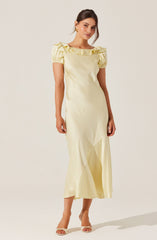 A woman is wearing a light yellow, full-length dress with short, puffy sleeves that have ruffle details. The dress has a round neckline with ruffled embellishments along the collar. She is also wearing open-toed, high-heeled sandals in a light color.