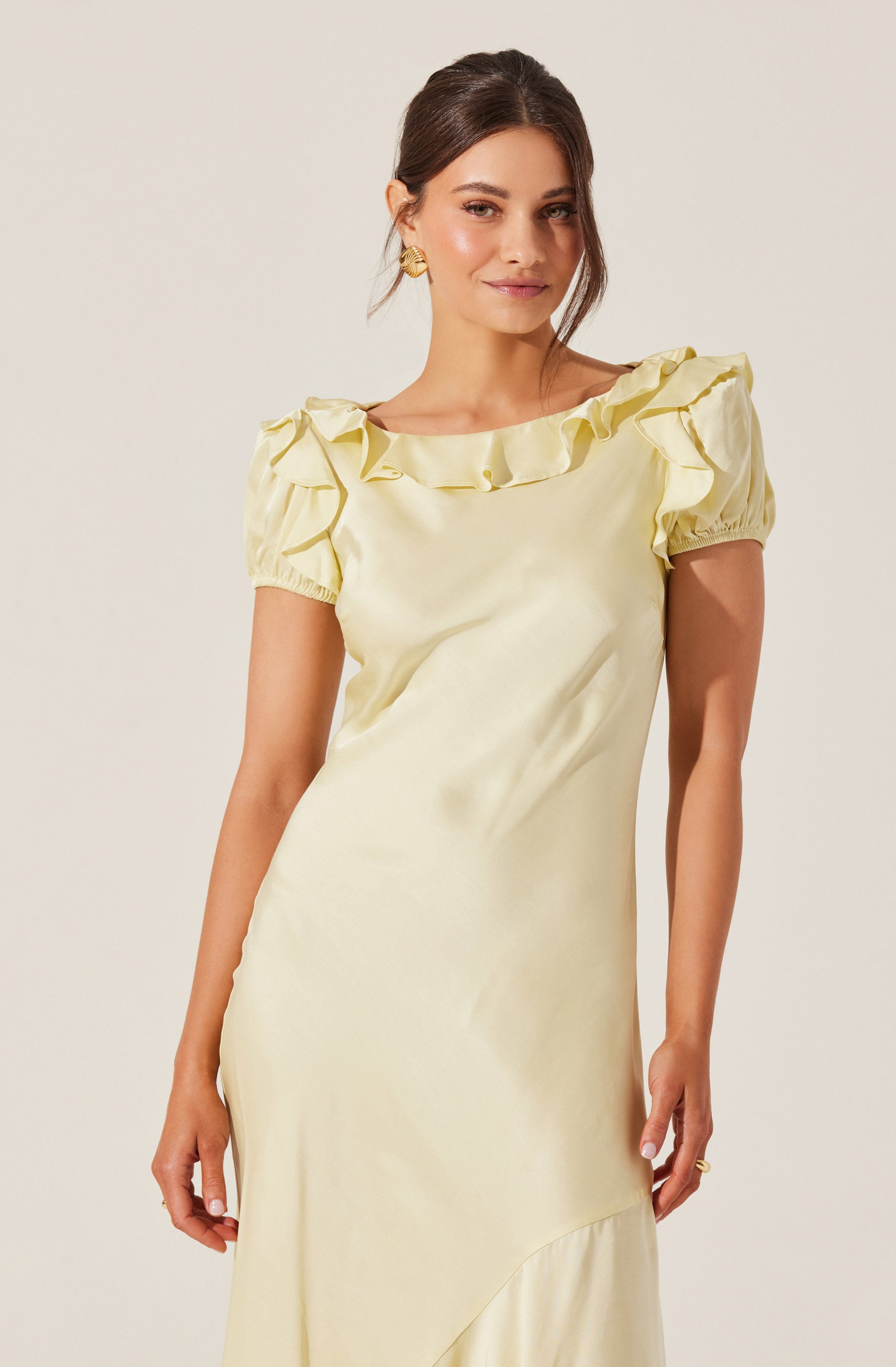 A woman is wearing an elegant cream-colored satin dress with a round neckline and short, puffed sleeves. The dress features ruffled details along the neckline. She has dark brown hair styled in a loose updo, and is accessorized with gold earrings.