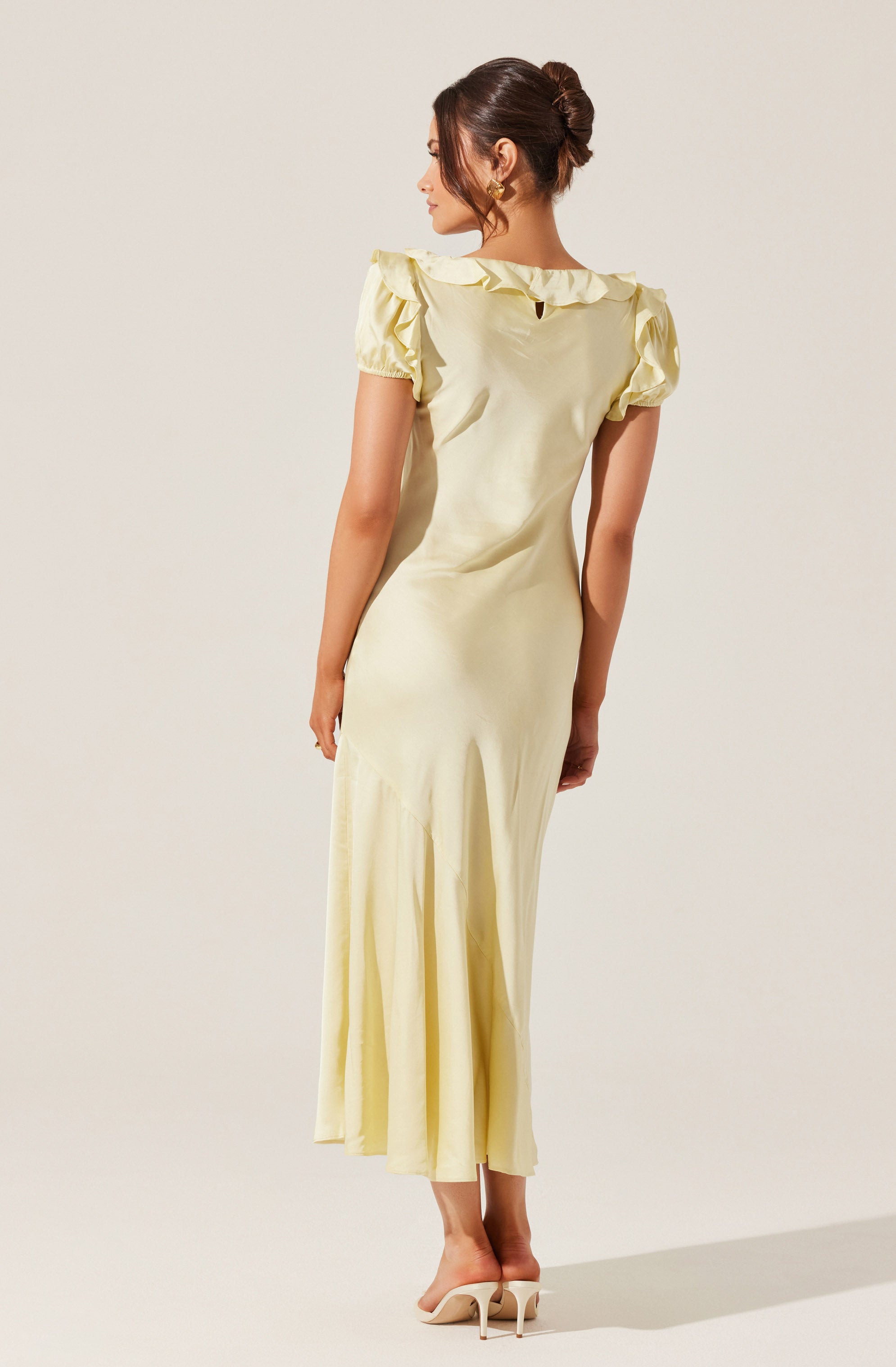 A woman is wearing a long, pale yellow dress with short puffed sleeves and ruffled detailing along the neckline. The dress has a fitted bodice and a flowing skirt that reaches the floor. She is also wearing nude high-heeled shoes.