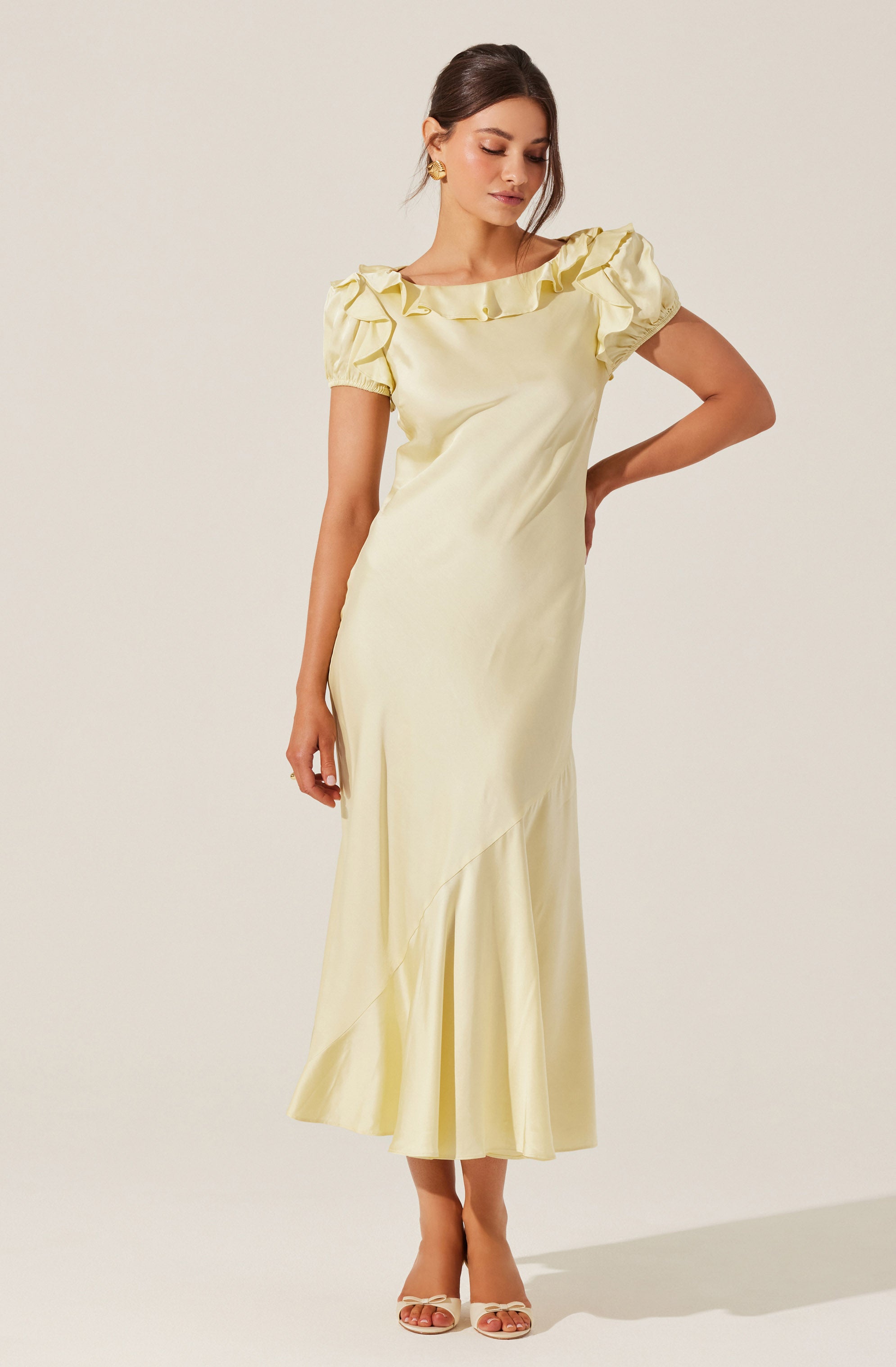 A woman is wearing a long, cream-colored satin dress with short puffed sleeves. The dress features a ruffled neckline and an asymmetrical hemline with a subtle pleated detail near the bottom. She is also wearing gold earrings and nude high-heeled sandals.