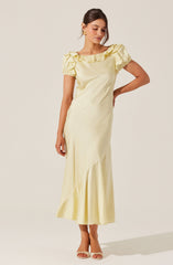 A woman is wearing a long, cream-colored satin dress with short puffed sleeves. The dress features a ruffled neckline and an asymmetrical hemline with a subtle pleated detail near the bottom. She is also wearing gold earrings and nude high-heeled sandals.