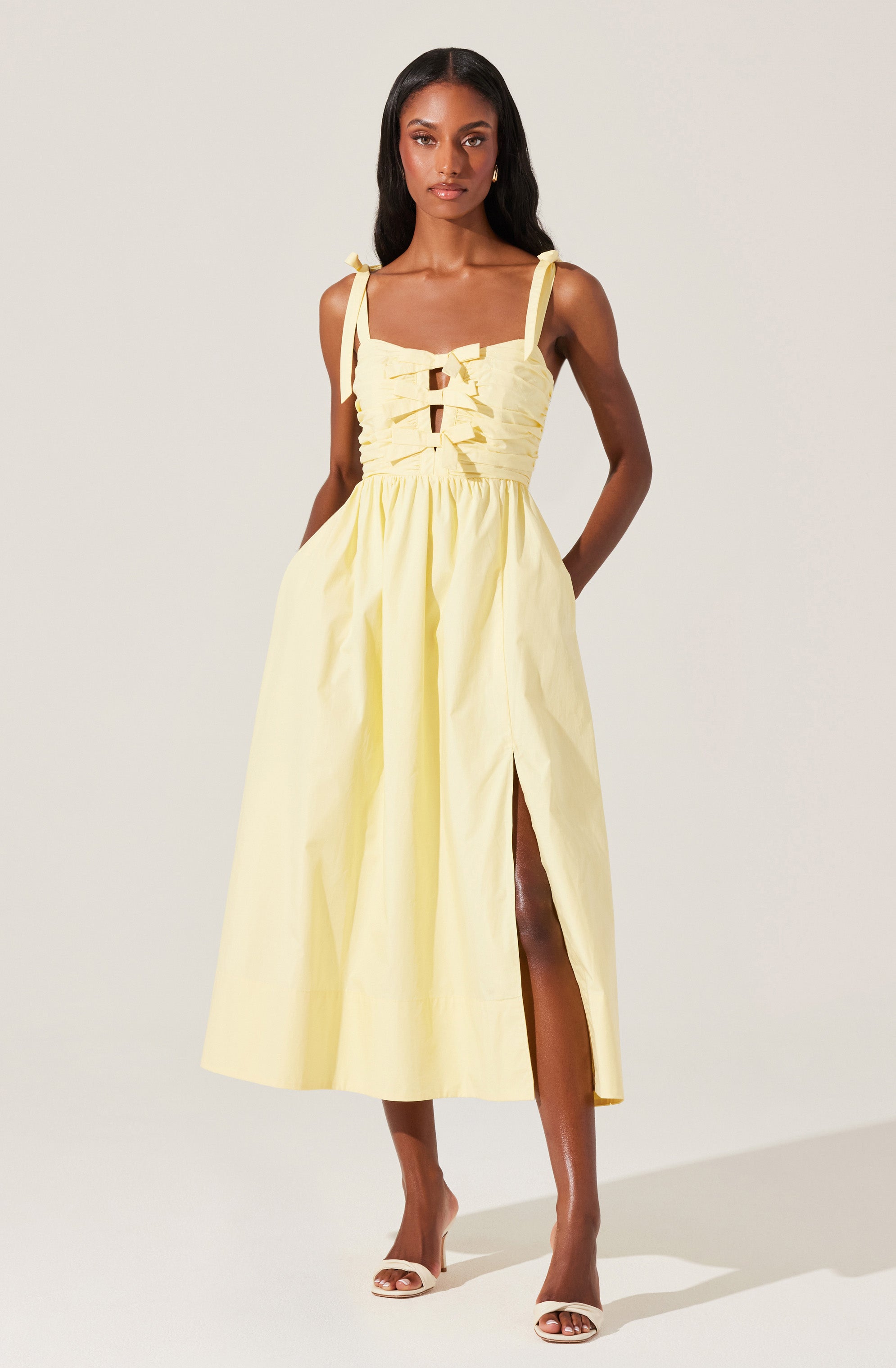A woman is wearing a sleeveless, light yellow midi dress with shoulder tie straps. The bodice features a ruched design with multiple horizontal gathers and a front tie detail. The skirt has a high slit on the left side, revealing part of her leg. She is also wearing white open-toe high heels.