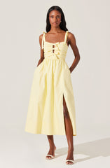 A woman is wearing a sleeveless, light yellow midi dress with shoulder tie straps. The bodice features a ruched design with multiple horizontal gathers and a front tie detail. The skirt has a high slit on the left side, revealing part of her leg. She is also wearing white open-toe high heels.