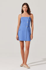 A woman is wearing a sleeveless, blue dress with thin spaghetti straps. The dress features an asymmetrical button-down design with three large buttons on the front and two decorative flap pockets on the hips. She is also wearing cream-colored, woven slip-on shoes.