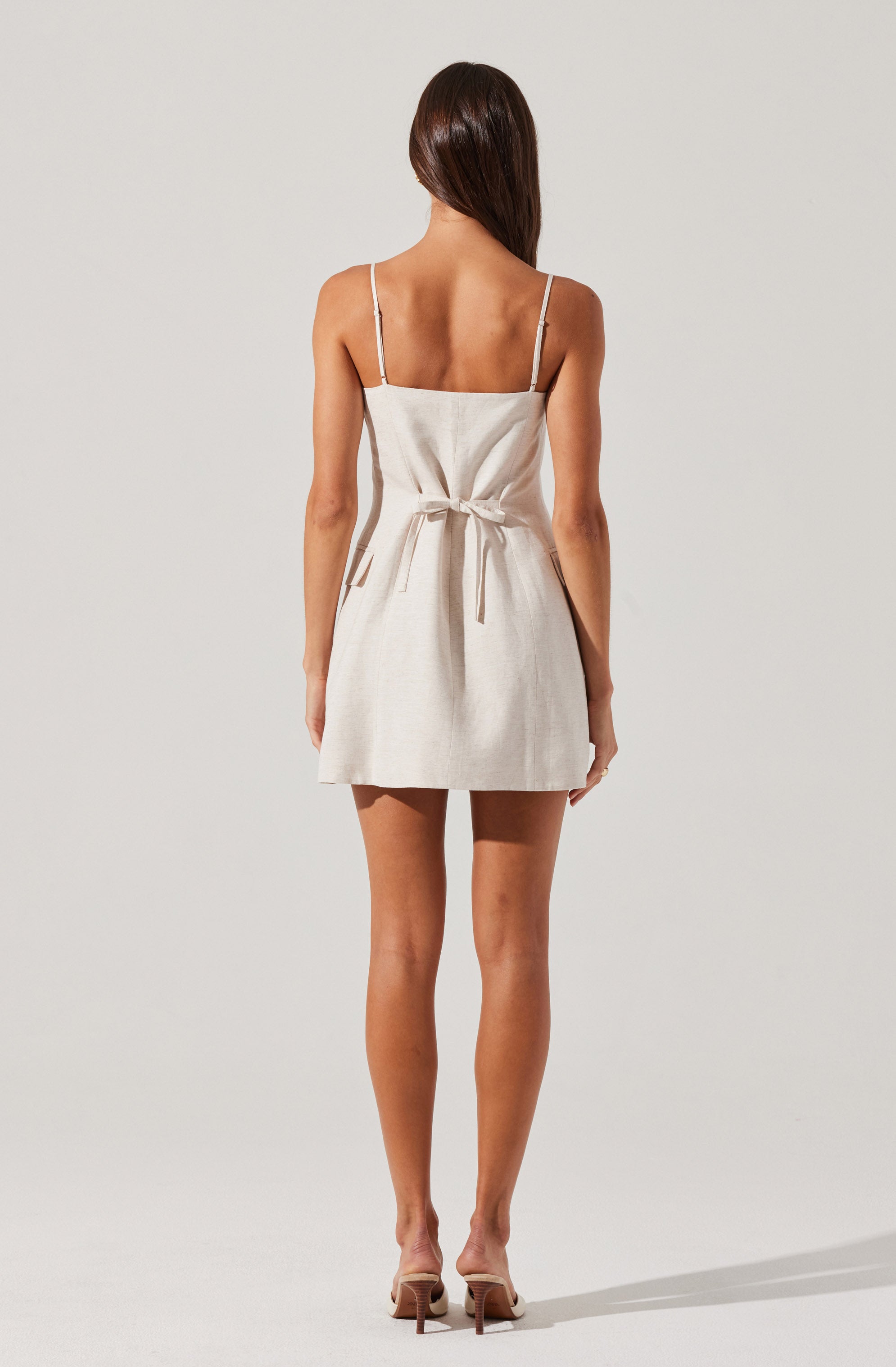 A woman is wearing a sleeveless dress with thin spaghetti straps. The dress is light-colored, possibly beige or off-white, and has a fabric belt tied at the waist. The hemline falls above the knees. She is also wearing nude-colored high-heeled shoes.