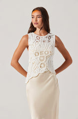 The woman is wearing a sleeveless, white crochet top with intricate circular and swirling patterns. She has paired it with a smooth, satin-like cream-colored skirt.