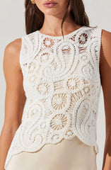 A woman is wearing a sleeveless white crochet top with intricate circular and spiral patterns throughout. The top has a round neckline and features a scalloped hem at the bottom.