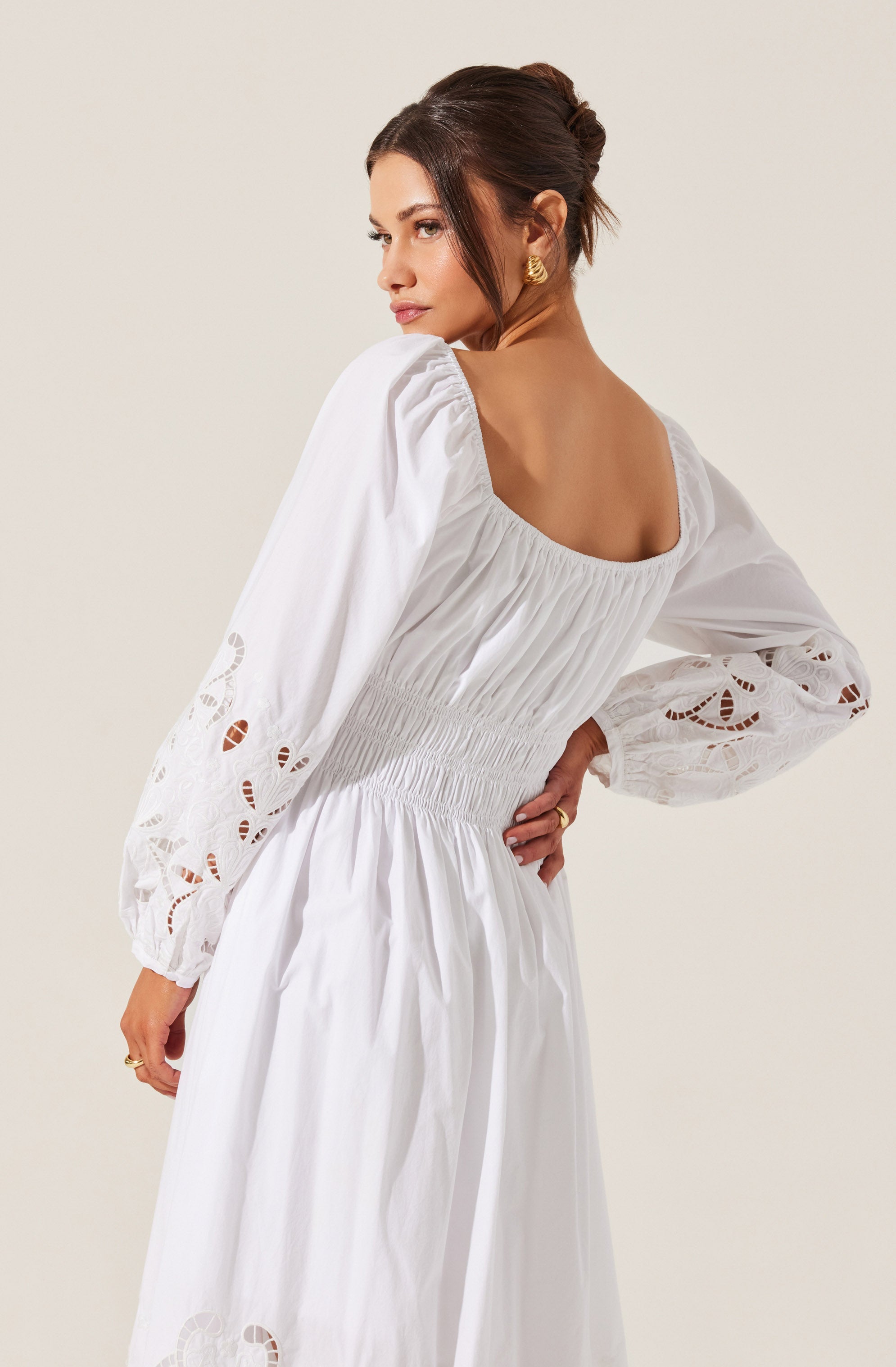 A woman is wearing a long white dress with a square neckline. The dress features puffed long sleeves with embroidered cutout details. The waist of the dress is cinched with gathered, elasticized smocking, and the fabric appears lightweight and airy. She has brunette hair styled in an elegant updo and is wearing gold earrings.