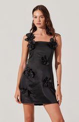 A woman is wearing a black satin mini dress adorned with large 3D floral embellishments. The dress features thin shoulder straps, with some flowers positioned on the straps and others scattered across the front of the dress. The floral decorations are made from the same satin material as the dress, creating a textured, three-dimensional effect.