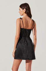 A woman is wearing a black satin dress with spaghetti straps that tie at the shoulders. The dress features a fitted bodice with a sheer panel at the back and a gathered waist, falling to a short hemline above the knees. She has dark brown hair and is accessorizing with gold earrings.