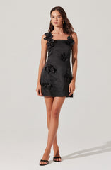 A woman is wearing a black sleeveless mini dress with thin spaghetti straps. The dress features large, decorative fabric flowers attached to the straps on the shoulders and scattered across the front of the dress. She pairs the dress with black high-heeled sandals and gold earrings.