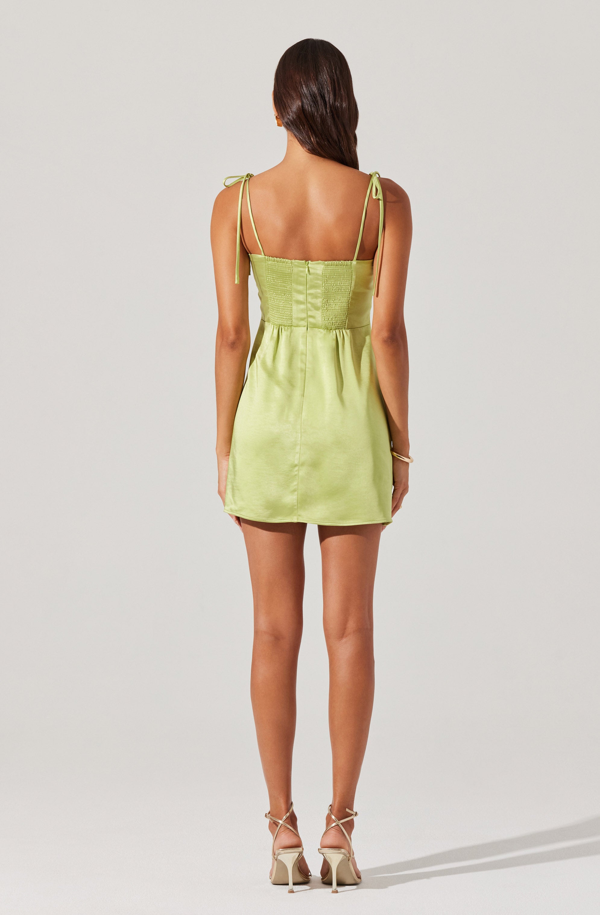 A woman is wearing a sleeveless, light green dress with thin, tie-up shoulder straps. The dress has a fitted bodice with a textured waistband and a slightly gathered skirt that ends above the knees. She is also wearing beige high-heeled shoes with ankle straps.