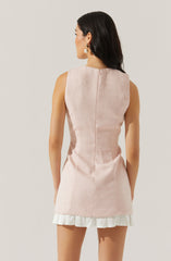 A woman is wearing a sleeveless, light pink textured dress with a straight fit. The dress has a hidden back zipper closure and ends above the knee. The hemline features a white ruffled trim. The woman is accessorized with large pearl drop earrings and has long, dark hair styled in loose waves.