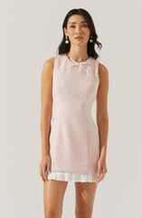 A woman is wearing a sleeveless, light pink textured dress with a small bow at the neckline. The dress is fitted and has a white ruffled hem at the bottom. She is accessorized with large, round earrings and has long, dark hair styled in loose waves.