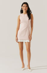 The woman is wearing a sleeveless, light pink dress with a subtle textured pattern. The dress has a small bow detail at the neckline and a white pleated ruffle hem at the bottom. She is also wearing white pointed-toe heels with a bow detail and pearl drop earrings.